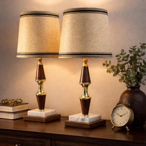May include: Two matching table lamps with beige fabric shades and black trim. The lamps have a dark wood and gold base, with a marble and wood base. A small gold alarm clock and a vase with greenery are also on the table.
