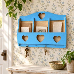 May include: A bright blue wooden wall-mounted organizer featuring three storage compartments, three heart-shaped cutouts, and four metal hooks. The unit holds various envelopes and papers, with two sets of metal keys hanging from the hooks below.