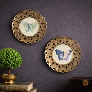 May include: Two decorative wall art pieces with gold-colored, ornate frames. Each frame holds a circular image of a butterfly. One butterfly is blue and green, the other is blue and pink. The background is a dark gray.
