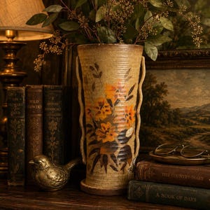 May include: A tall, tan ceramic vase with orange floral designs and two curved handles. The vase holds leafy branches. Antique books and a brass bird figurine sit nearby, creating a vintage aesthetic. A lamp and framed artwork are in the background.
