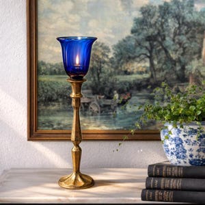 May include: A tall, antique-style candle holder with a vibrant blue glass cup holding a lit candle. The candle holder is gold-toned, with a detailed, textured design. The background features a framed landscape painting and a potted plant.