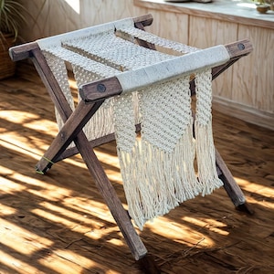 May include: A wooden luggage rack with a macrame top and fringe. The frame is made of dark brown wood, and the macrame is a light beige color. The rack is folded and has a rectangular shape.