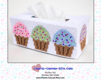 May include: A white plastic canvas tissue box cover with a design of three cupcakes. The cupcakes are decorated with pink, green, and blue frosting and colorful sprinkles. The tissue box cover is labeled "0645 Cupcakes Long Tissue Topper" and "Plastic-Canvas-Kits.com".