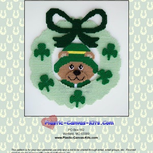 May include: A green plastic canvas wreath with a green shamrock design and a green and yellow bear wearing a green hat. The wreath has a green bow on top. The text "Plastic-Canvas-Kits.com" is at the bottom of the image.