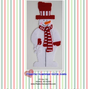 Op de afbeelding: Rood en wit plastic canvas sneeuwman wanddecoratie patroon. De sneeuwman draagt een rode hoed met witte strepen, een rood-wit gestreepte sjaal en rode wanten. Het patroon is van Plastic-Canvas-Kits.com.