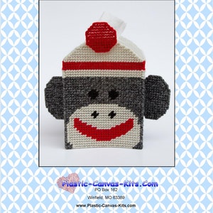 May include: A plastic canvas pattern for a sock monkey tissue box cover. The pattern features a gray monkey face with a red smile and a white hat with a red pom-pom. The pattern is titled "0516 Sock Monkey Tissue Topper" and includes the website address "Plastic-Canvas-Kits.Com".