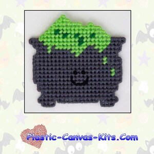 May include: A gray and green plastic canvas pattern of a witch's cauldron with a smiling face. The pattern is for a magnet and is titled "2002 Witch's Cauldron Magnet".