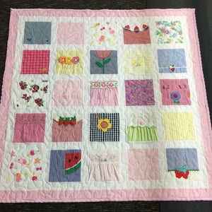 May include: A square patchwork quilt featuring a pink border and a white background with quilted patterns. The quilt consists of twenty-five individual squares, each displaying various fabric textures, floral prints, gingham patterns, and appliqu&eacute;d motifs.