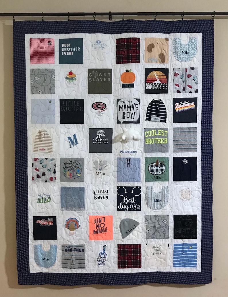 May include: A memory quilt constructed from various fabric squares featuring printed text and graphics, including phrases like Best Brother Ever, Giant Slayer, Mama's Boy, and Coolest Brother. The quilt has a dark blue border and white stitching.