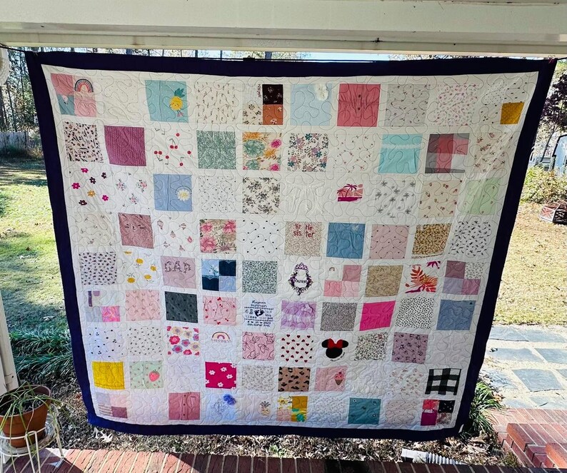 May include: A memory quilt composed of numerous square fabric patches featuring various patterns, colors, and embroidered designs, including a Minnie Mouse icon, a crown, and the text GAP, all framed by a solid dark blue border with white stitching.