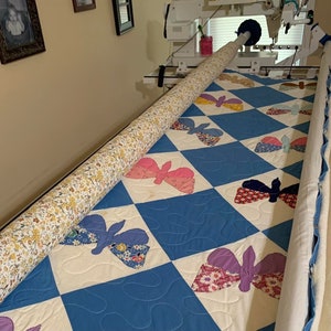 May include: A close-up of a quilt with a blue and white diamond pattern. The quilt is being rolled up on a quilting machine. The quilt features a variety of colorful butterfly designs.