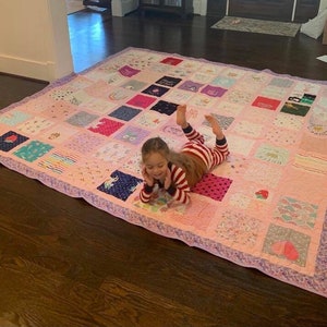 May include: A large square patchwork quilt constructed from various fabric squares featuring diverse patterns, colors, and graphic prints. The quilt is bordered by a light purple patterned fabric and displays a grid of colorful textile blocks.