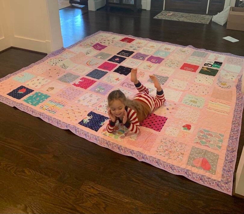 May include: A large square patchwork quilt constructed from various fabric squares featuring diverse patterns, colors, and graphic prints. The quilt is bordered by a light purple patterned fabric and displays a grid of colorful textile blocks.