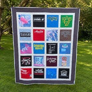 May include: A memory quilt featuring a grid of twenty square panels made from various graphic t-shirts. The quilt has a dark gray border and white sashing between the squares. The panels display various theatrical and pop culture logos and text.