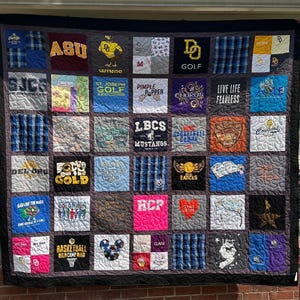 May include: A memory quilt composed of various square fabric patches featuring diverse graphic prints, logos, and text including ASU, St. Joseph Golf, LBCS Mustangs, and Live Life Fearless. The quilt has a dark border and visible quilted stitching.