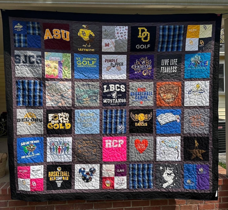 May include: A memory quilt composed of various square fabric patches featuring diverse graphic prints, logos, and text including ASU, St. Joseph Golf, LBCS Mustangs, and Live Life Fearless. The quilt has a dark border and visible quilted stitching.