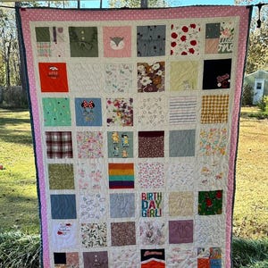 May include: A memory quilt featuring a grid of assorted fabric squares with various patterns, colors, and graphic prints, including text that reads "BIRTH DAY GIRL!" and "Little Broncos Fan." The quilt is bordered by a pink and white polka dot fabric.