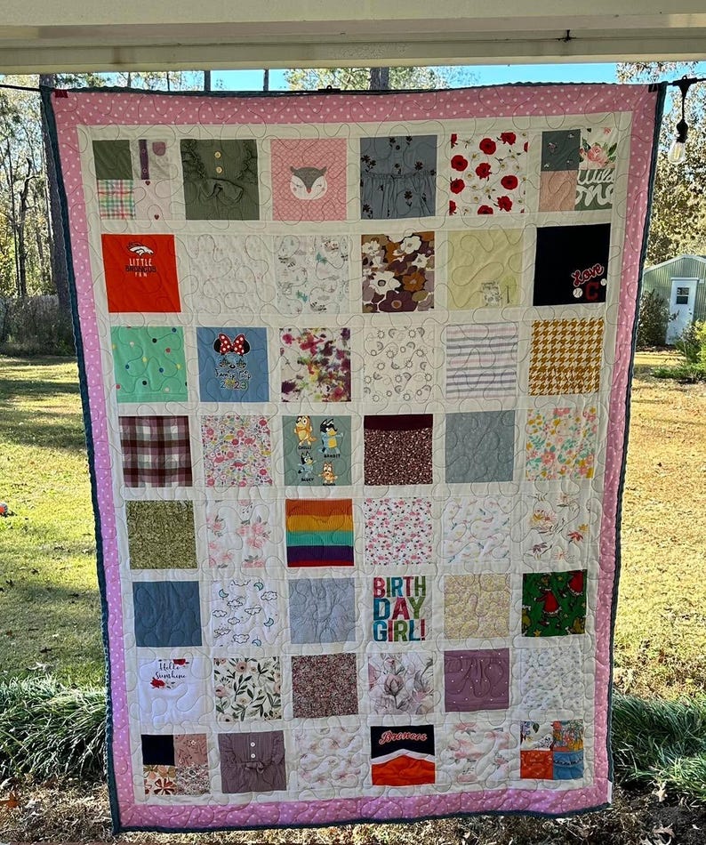 May include: A memory quilt featuring a grid of assorted fabric squares with various patterns, colors, and graphic prints, including text that reads "BIRTH DAY GIRL!" and "Little Broncos Fan." The quilt is bordered by a pink and white polka dot fabric.