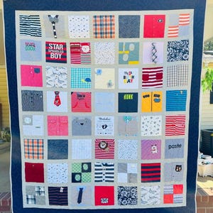 May include: A memory quilt composed of numerous square fabric patches featuring various patterns, colors, and graphic prints from children's clothing. The quilt has a dark blue border with a swirling stitch pattern and a light gray inner border.