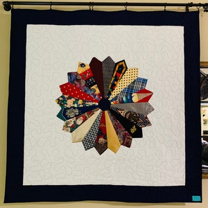 May include: A square quilt featuring a central circular medallion made of various neckties arranged in a sunburst pattern. The quilt has a white background with a continuous meandering stitch pattern and a solid dark navy blue border frame.