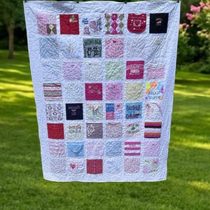 May include: A memory quilt constructed from various square fabric patches in shades of pink, white, red, and blue. The quilt features diverse embroidered graphics, text, and patterns, all stitched together with a white quilted background.