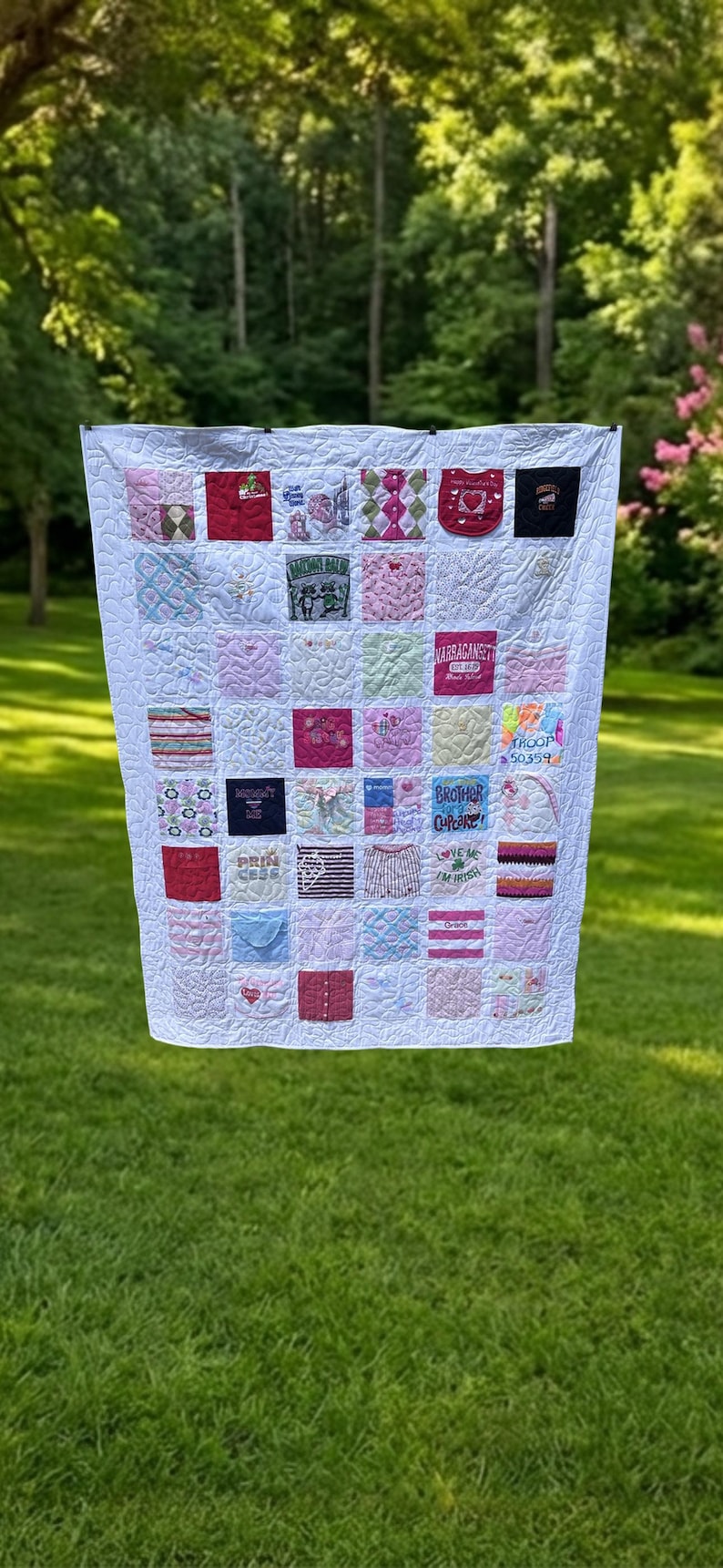 May include: A memory quilt constructed from various square fabric patches in shades of pink, white, red, and blue. The quilt features diverse embroidered graphics, text, and patterns, all stitched together with a white quilted background.