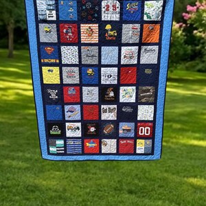 May include: A memory quilt constructed from various square fabric patches featuring diverse graphic prints, embroidered text, and sports themes. The quilt has a dark blue border and a light blue inner frame, with a grid layout of colorful squares.