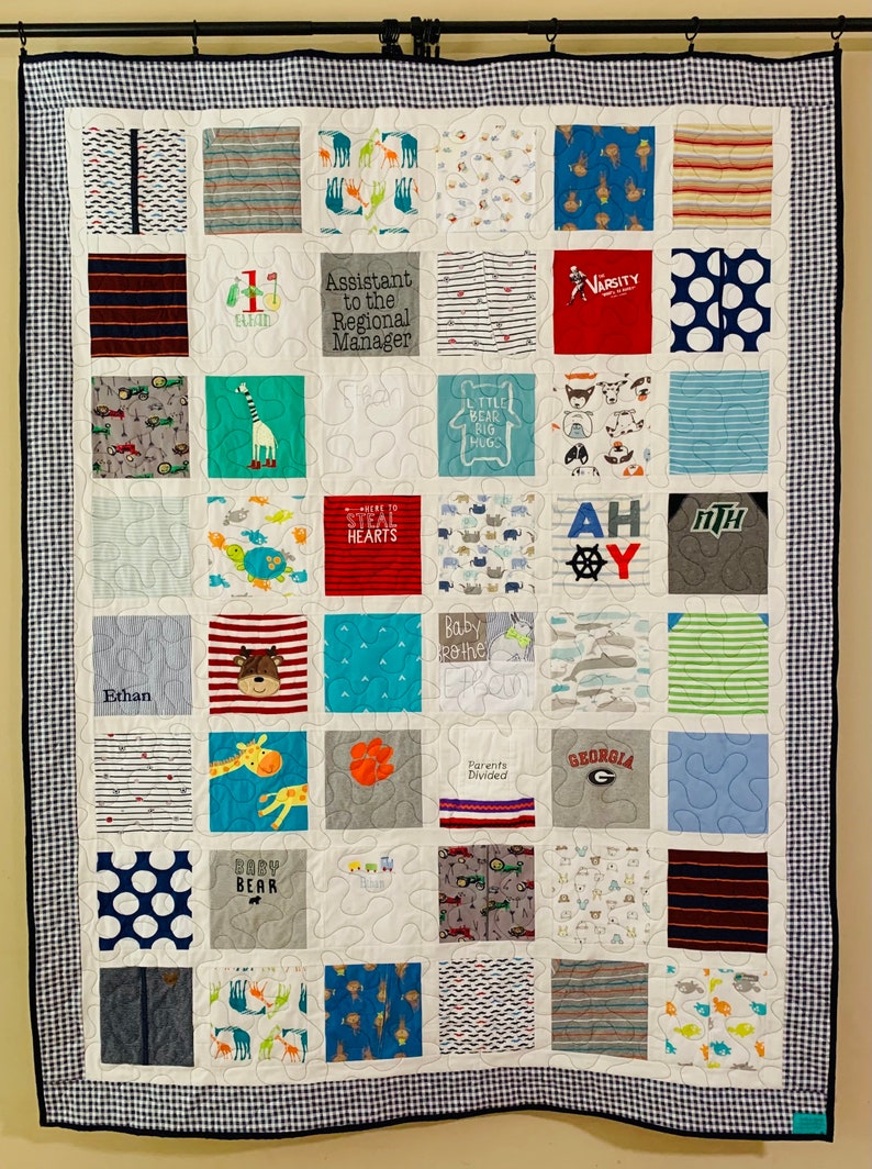May include: A memory quilt composed of various fabric squares featuring diverse patterns, animal prints, and text including Assistant to the Regional Manager, Varsity, Ahoy, Baby Bear, and Georgia. The quilt has a blue and white gingham border.
