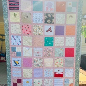 May include: A memory quilt composed of various fabric squares featuring diverse patterns, sports logos, and baby-themed prints. A central square displays birth details for Kennedy, including 7 lbs, 20.5 in, and 3 17 2025. The quilt is framed by a pink border and a green floral outer edge.