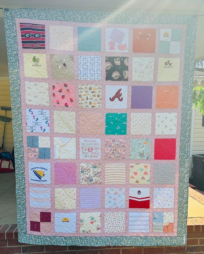 May include: A memory quilt composed of various fabric squares featuring diverse patterns, sports logos, and baby-themed prints. A central square displays birth details for Kennedy, including 7 lbs, 20.5 in, and 3 17 2025. The quilt is framed by a pink border and a green floral outer edge.