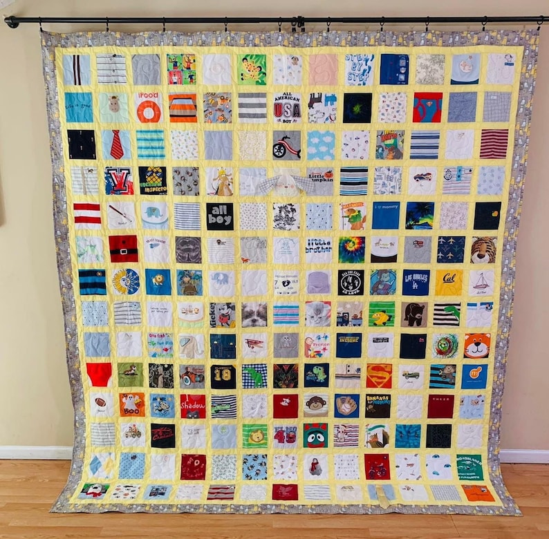 May include: A large memory quilt composed of numerous square fabric patches featuring various graphic prints, patterns, and text including "All American Boy," "Little Brother," and "Los Angeles." The quilt has a yellow grid and a gray border.