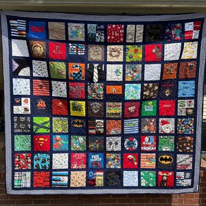 May include: A large memory quilt composed of numerous square fabric patches featuring various colorful patterns, cartoon characters, and graphic prints. The quilt is framed by a dark blue border with a white and blue checkered edge trim.