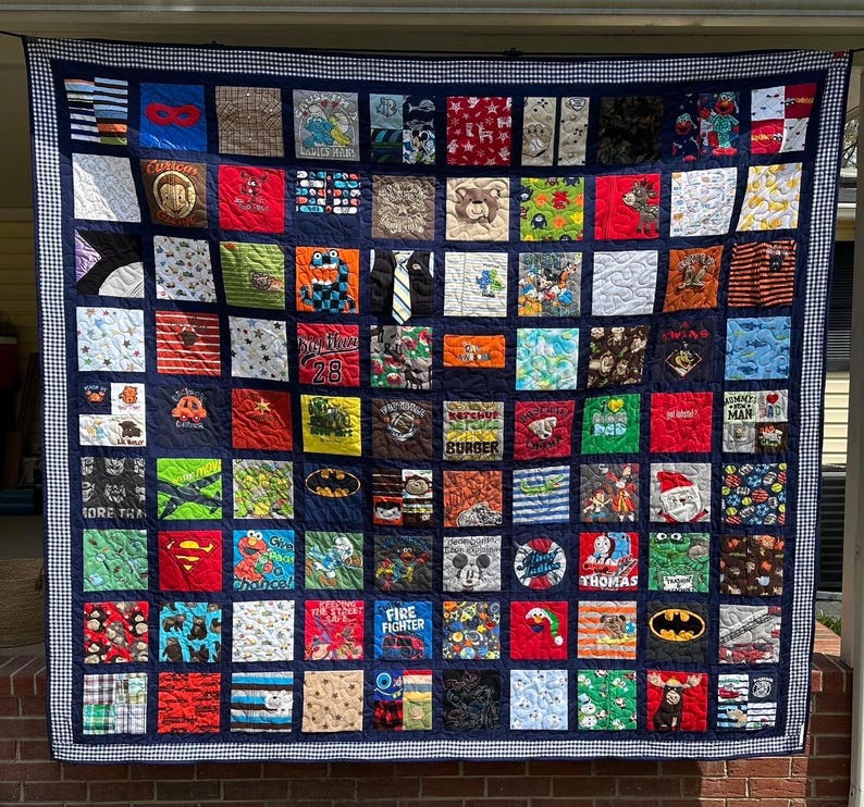 May include: A large memory quilt composed of numerous square fabric patches featuring various colorful patterns, cartoon characters, and graphic prints. The quilt is framed by a dark blue border with a white and blue checkered edge trim.
