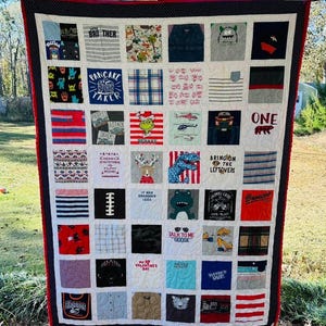 May include: A memory quilt constructed from various fabric squares featuring diverse graphic prints, text, and patterns. The quilt is bordered by a dark blue fabric with white polka dots and a thin red trim, featuring a white grid stitching pattern.