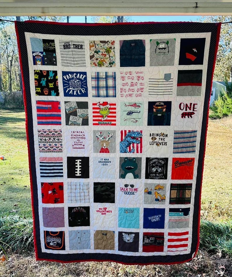 May include: A memory quilt constructed from various fabric squares featuring diverse graphic prints, text, and patterns. The quilt is bordered by a dark blue fabric with white polka dots and a thin red trim, featuring a white grid stitching pattern.