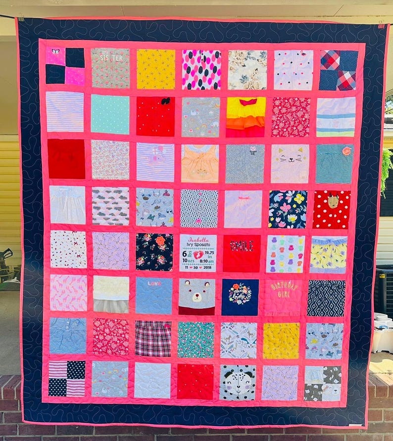 May include: A memory quilt composed of numerous square fabric patches featuring various patterns, colors, and graphics. A central white patch displays the text Isabella Ivy Sposato, 6 lbs 10 oz, 19.75 in, 8:10 am, 11-30-2018. The quilt has a bright pink border and a dark navy blue outer frame with a swirling stitched pattern.