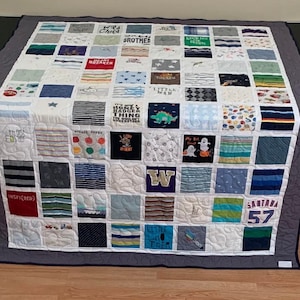 May include: A memory quilt constructed from a grid of assorted fabric squares featuring various colors, patterns, and printed text including "Wild Child," "Little Man," "Honey Badger," "My 1st Boat," "Inspired," and "Santana 57" with a dark gray border.