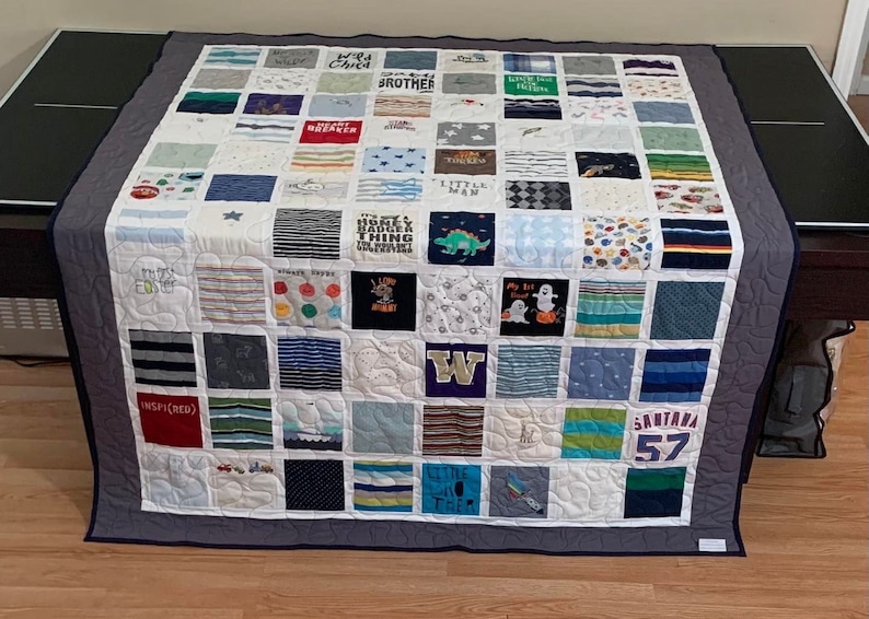 May include: A memory quilt constructed from a grid of assorted fabric squares featuring various colors, patterns, and printed text including "Wild Child," "Little Man," "Honey Badger," "My 1st Boat," "Inspired," and "Santana 57" with a dark gray border.