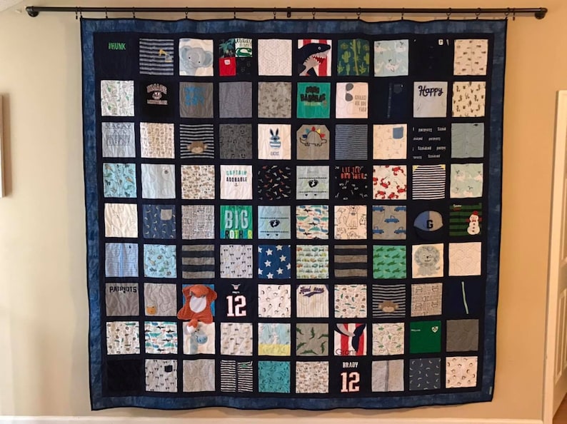 May include: A large square memory quilt composed of numerous fabric squares featuring various patterns, colors, and graphics, including text like "Big Brother," "Happy," and "Brady 12." The quilt is framed by a dark blue border and hangs from a black rod.