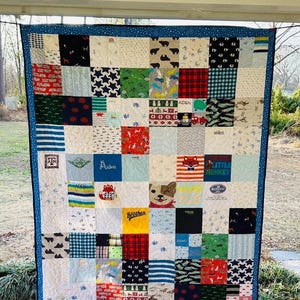 May include: A patchwork quilt constructed from various fabric squares featuring diverse patterns, colors, and graphics, including the names Aiden and Brother, a fox, a dog, and a star motif, all bordered by a dark blue fabric with a white star pattern.