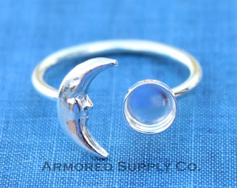 May include: Silver ring with a crescent moon design and a round bezel for a gemstone. The ring is on a blue fabric background. "ARMORED SUPPLY CO." is printed on the fabric.
