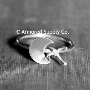 May include: A silver ring with a crescent moon and a star design. The ring is on a textured gray surface. © Armored Supply Co.