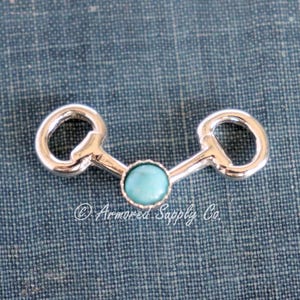May include: Silver-toned metal charm with a turquoise-colored stone center. The charm has a design reminiscent of a horse bit. The charm is set against a textured blue background.