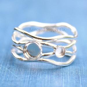 May include: Silver ring with three wavy bands and two round bezel settings. The ring is on a blue fabric background. Armored Supply Co. is printed on the ring.