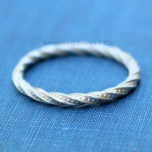 Op de afbeelding: Een zilveren ring met een gedraaid ontwerp en kleine kralen langs de rand. De ring ligt op een blauwe stoffen achtergrond.