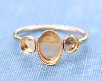 May include: Gold ring with three oval-shaped settings for adding stones or charms. The ring is on a blue fabric background.