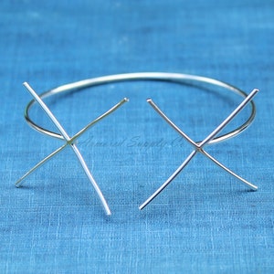 Puede incluir: Dos engastes de metal plateado para la elaboración de joyas. Los engastes tienen forma de cruz con cuatro garras. Los engastes están sobre un fondo de tela azul.