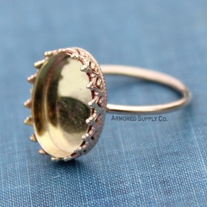 Op de afbeelding: Een ring van goudkleurig metaal met een decoratieve kroonvormige bezel. De bezel is ontworpen om een cabochon-steen of andere versiering te bevatten. De ring heeft een eenvoudige band.