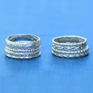 May include: Two silver rings with intricate designs are displayed against a blue background. The rings feature detailed patterns, including floral and braided textures. The rings are stacked, showcasing multiple bands with varying embellishments.