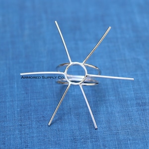May include: A silver-colored ring setting with six prongs extending outwards from a central circular design. The ring setting is displayed against a blue background. The text "ARMORED SUPPLY CO." is visible.